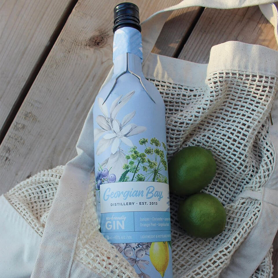 A bottle of gin in a re-usable bag with two limes sitting on wooden planks