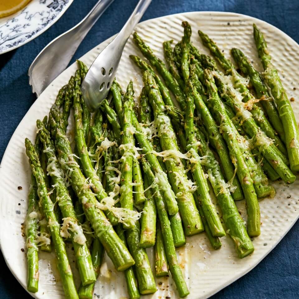 A white platter with asparagus topped with Parmesan cheese, with tongs next to it on a blue tablecloth