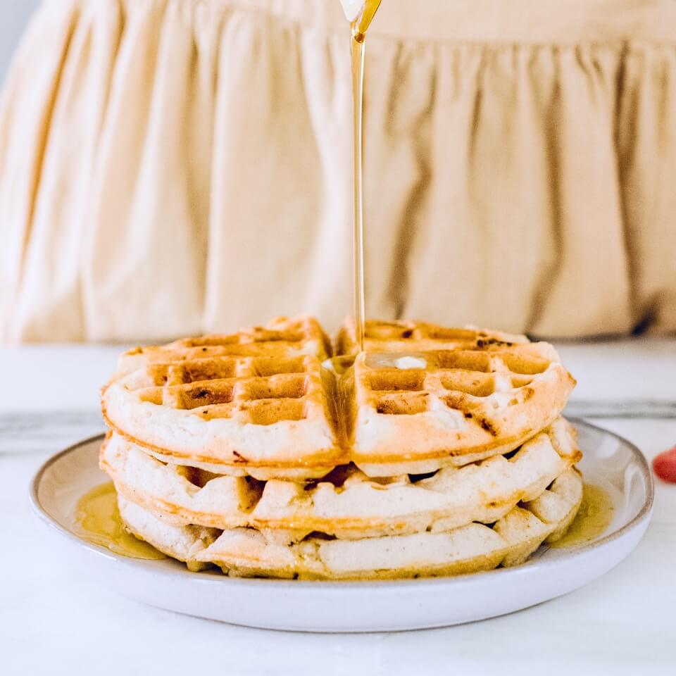 A plate of waffles with someone in a light orange shirt pouring syrup overtop