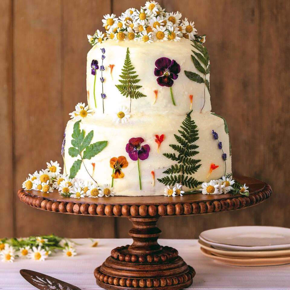 A two-tiered cake with white frosting and flowers and leaves pressed into it on a wooden cake stand, with a server, plates and a wooden panelled wall visible behind it