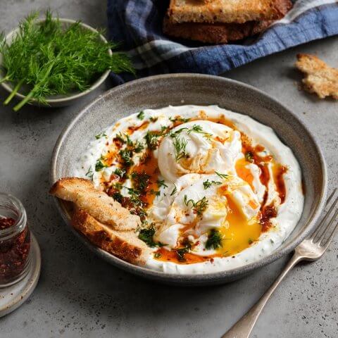 A grey dish with eggs and small toasts on a matching grey surface with garnishes, more bread and a fork also visible on the table