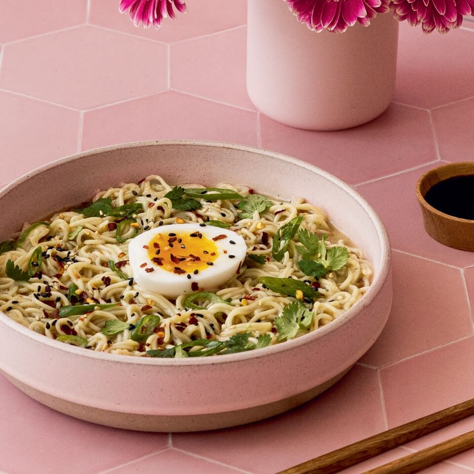 A pink bowl with ramen on a pink tiled surface with flowers visible in the background