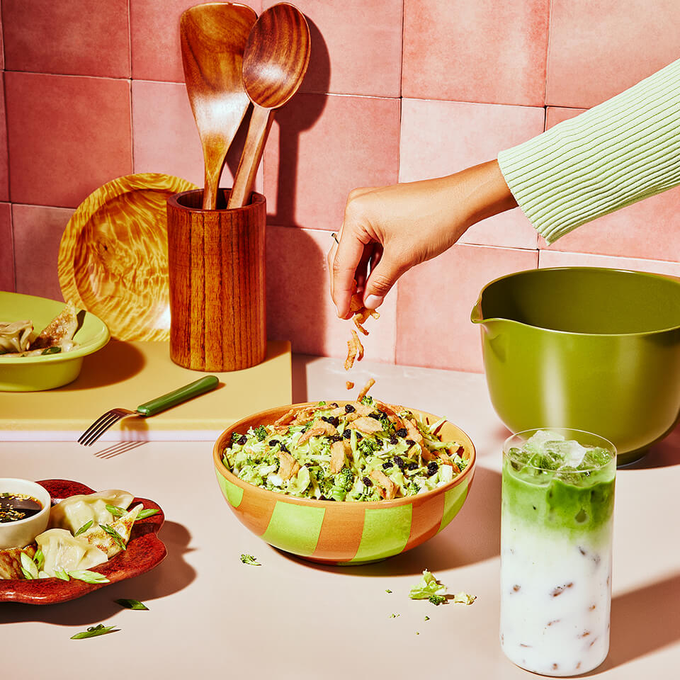 A person's arm sprinkling something on a salad in a bowl on a counter