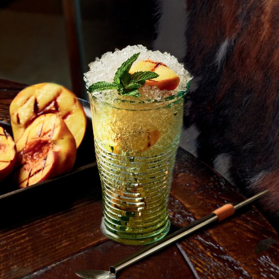 A mint julep cocktail with grilled peaches in a glass with more peaches and a bar spoon visible around it