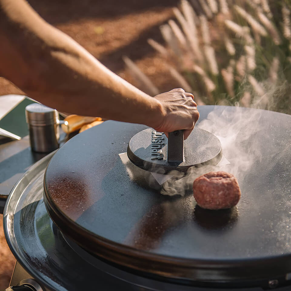 A person using a cast iron burger press on an outdoor griddle