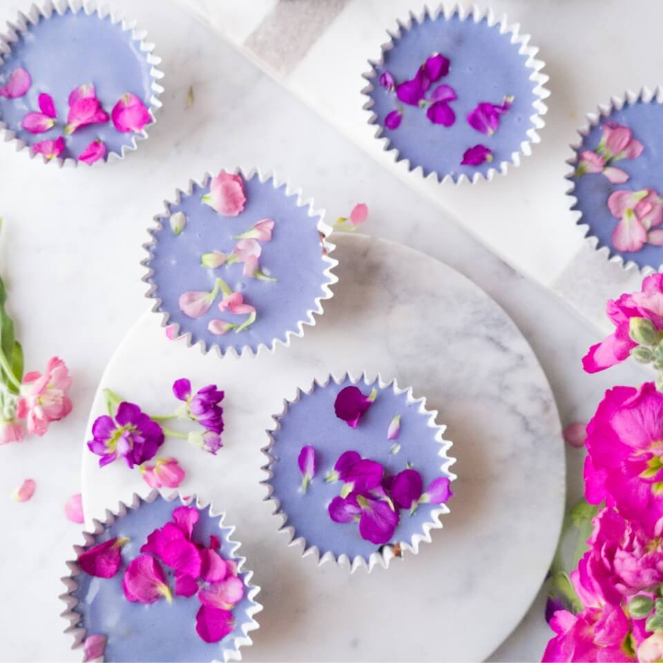 Purple-glazed brownies topped with edible flowers in cupcake wrappers on a marble surface