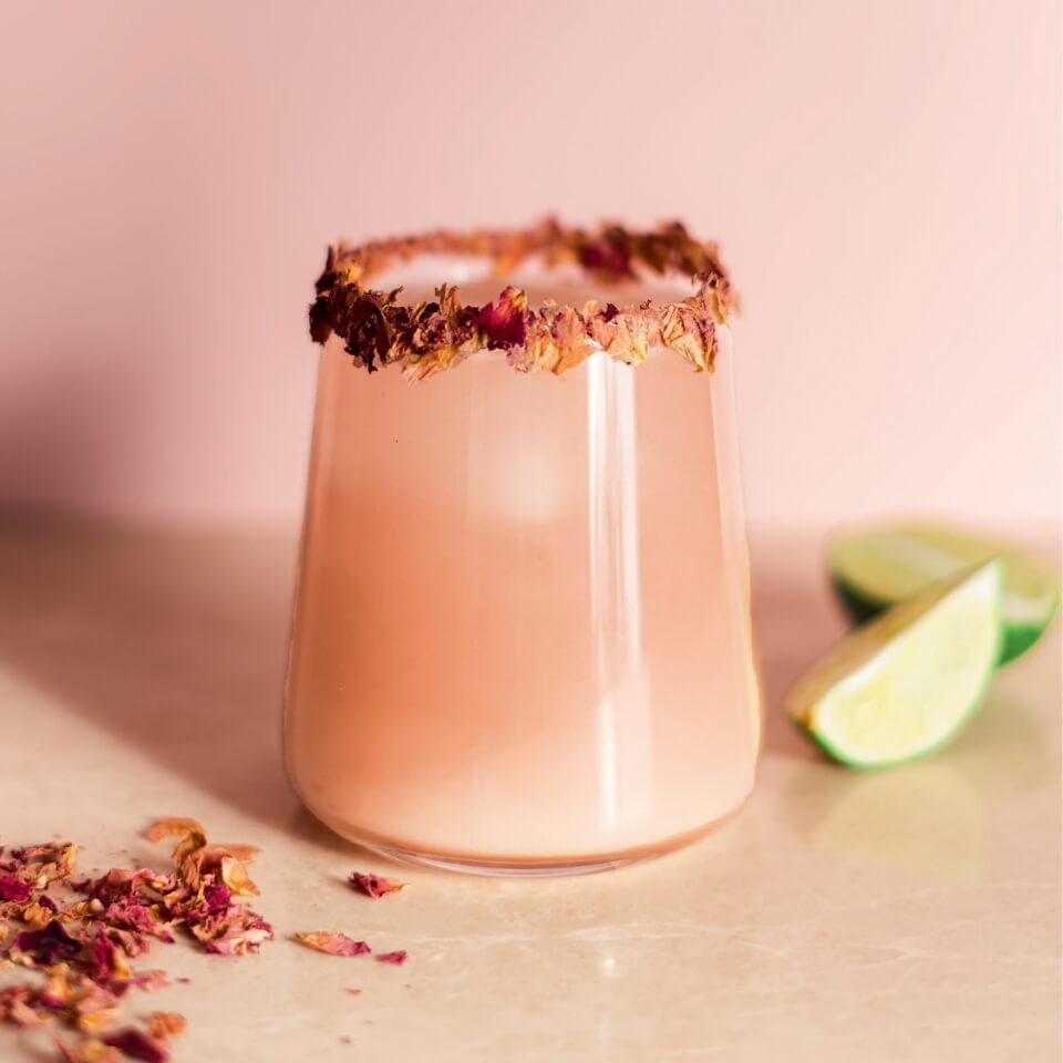 A glass with a pink drink and dried flowers on the rim, with lime wedges, more dried flowers, a tan countertop and pink wall visible in the background