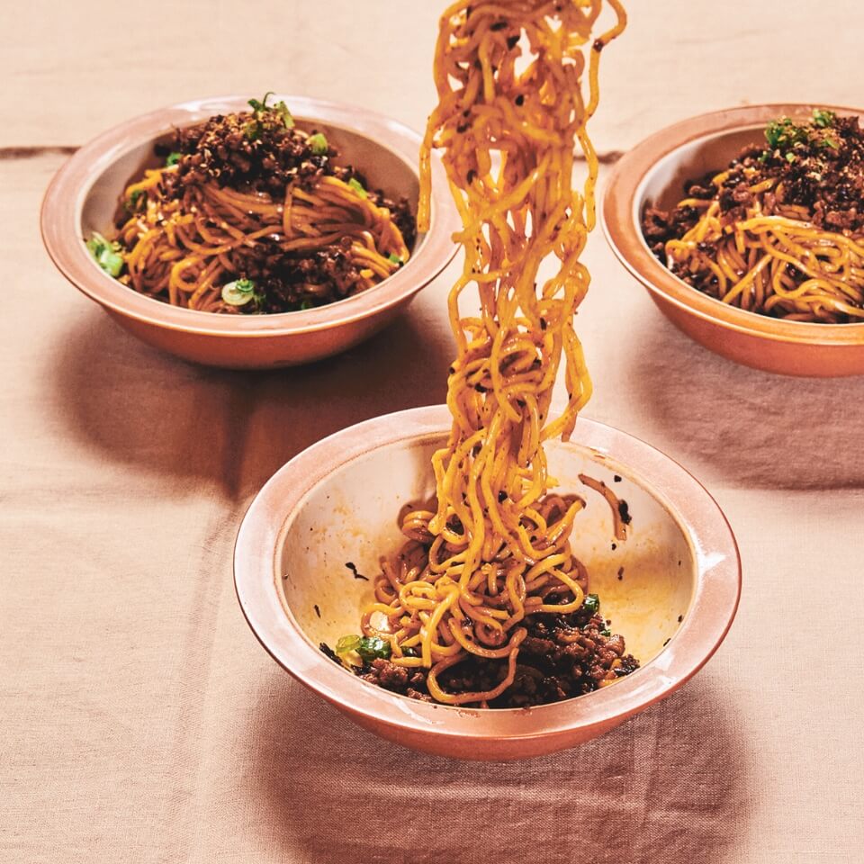 Three light pink bowls filled with noodles, one with them coming up out of the bowl, on a light pink tablecloth