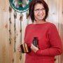 A woman in a red sweater, Jolene Johnson, holding maple syrup in front of a wooden wall with a dreamcatcher