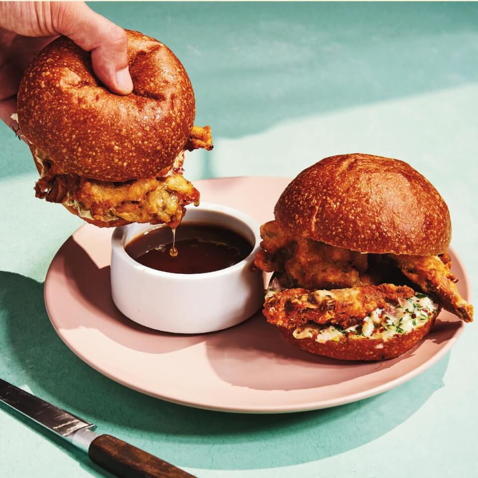 Two mushroom dip sandwichs and a person's hand dipping one on a light pink plate on a light blue surface