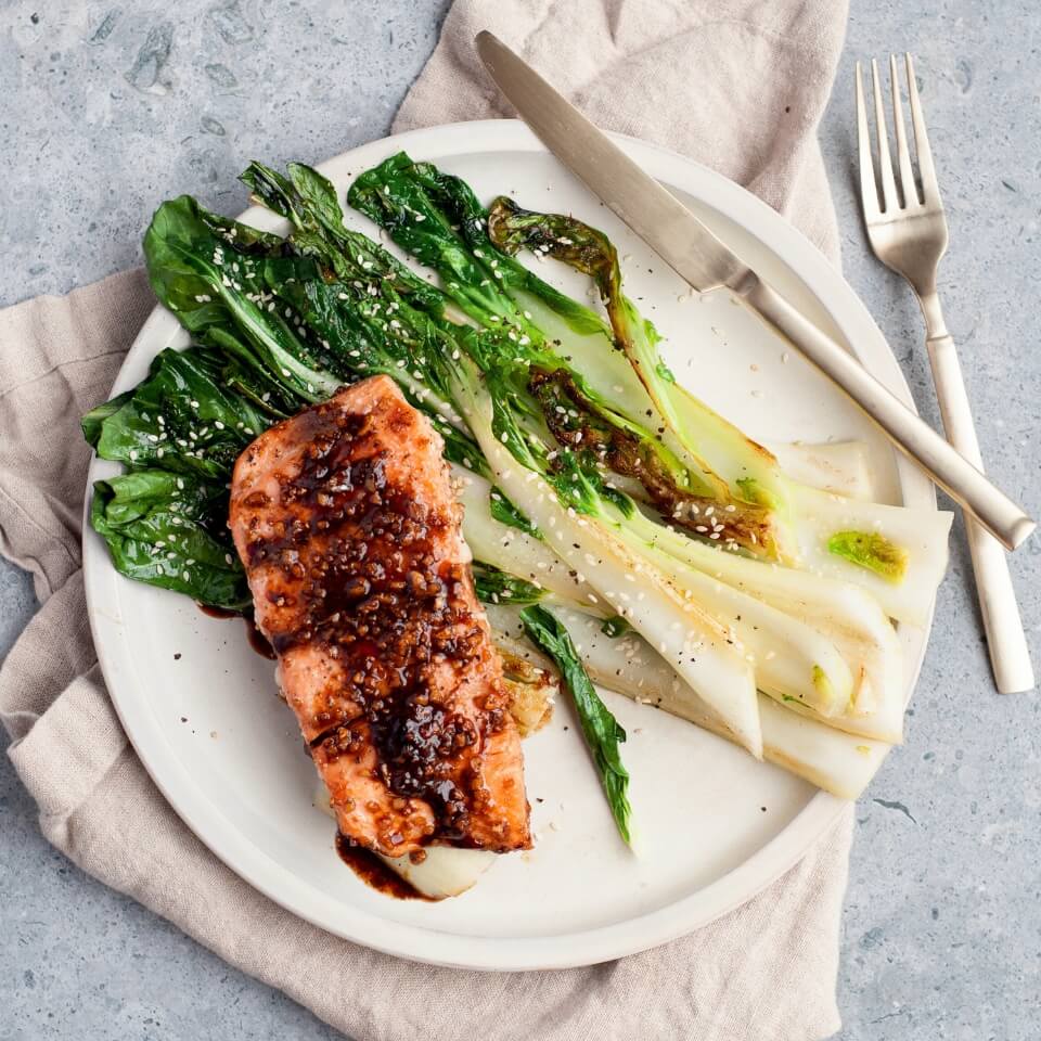 A plate with salmon and bok choy on a napkin with cutlery next to it