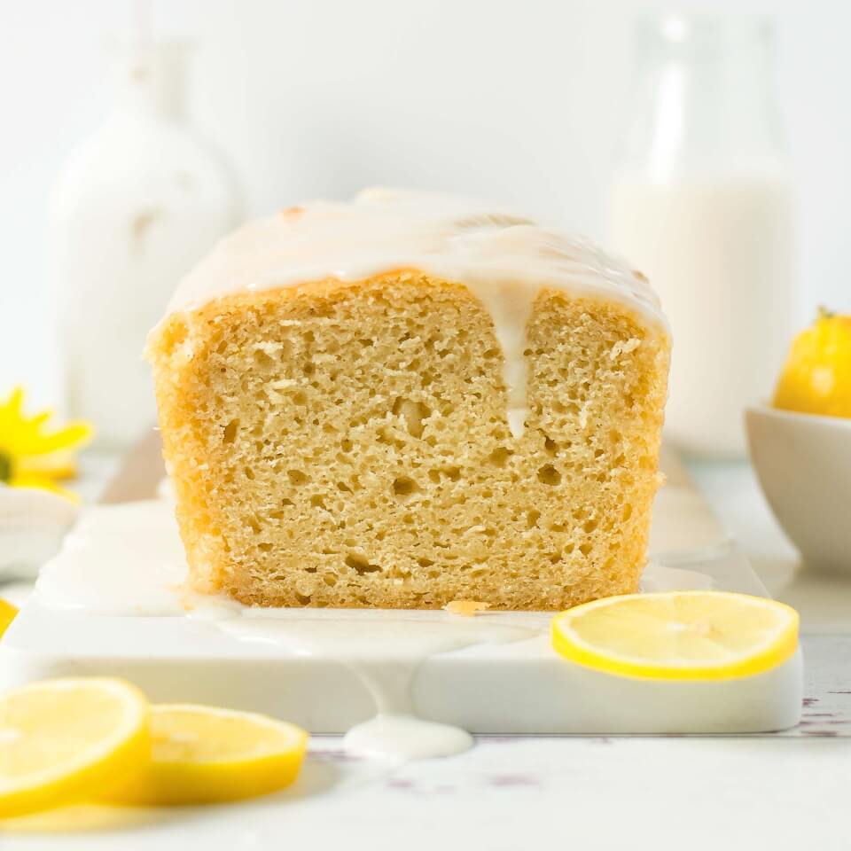 A frosted lemon loaf on a white surface with sliced lemons around it