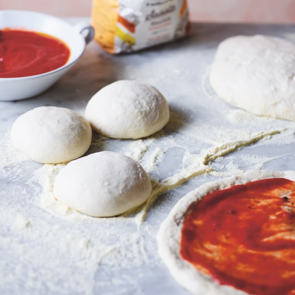 Balls of pizza dough, a bowl of sauce and flour, with one pizza partway through preparation