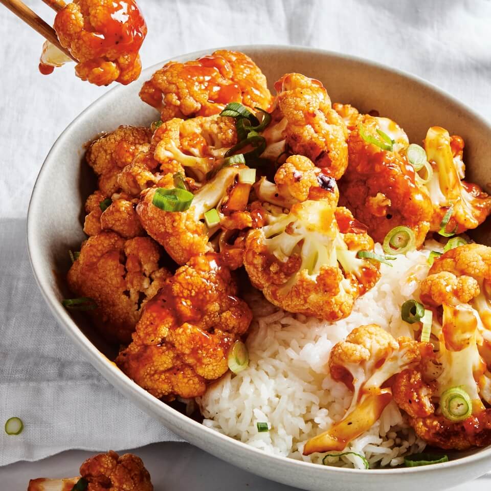 A bowl of cauliflower in orange-red sauce and rice with chopsticks lifting a piece out