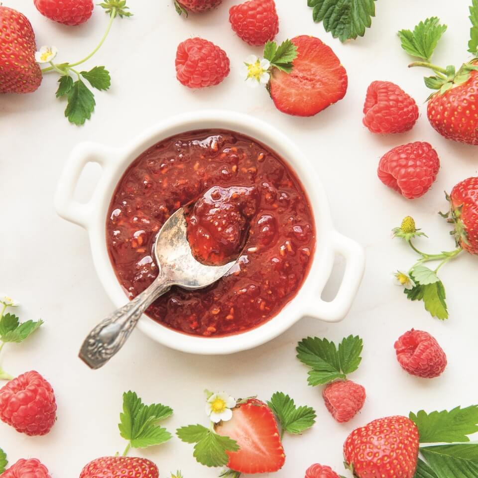 A white jar of jam with strawberries and raspberries around it