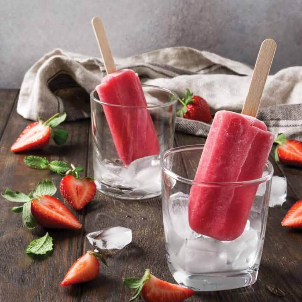 Strawberry popsicles in glasses over ice with sliced strawberries and a towel visible behind it