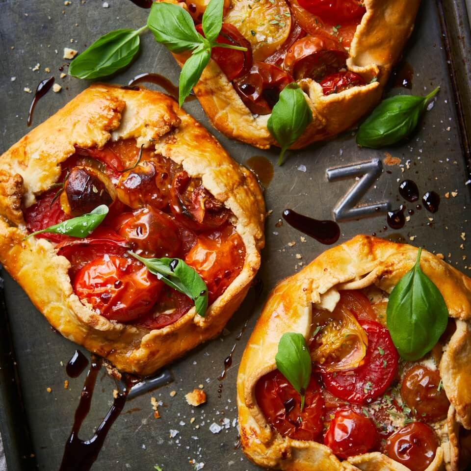 Miniature galettes with cherry tomatoes and a balsamic drizzle on a baking sheet