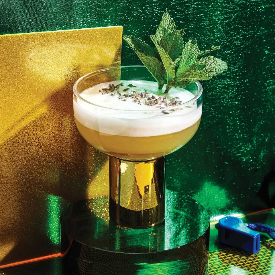 A cocktail i a coupe glass garnished with mint in front of a yellow-and-green backdrop