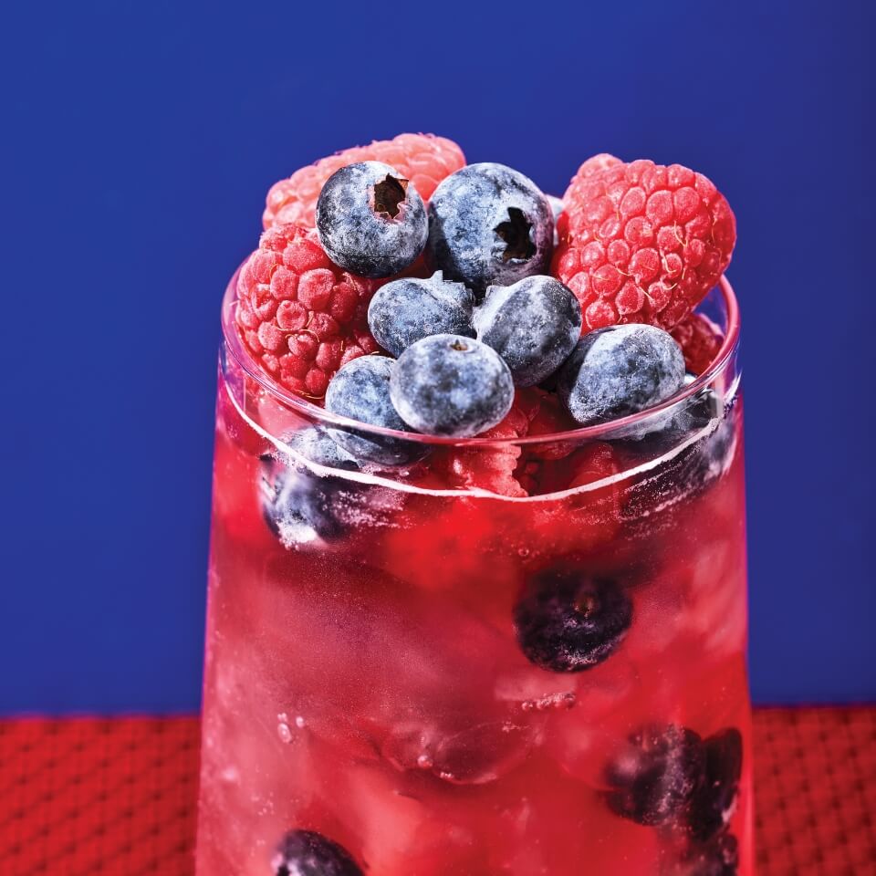 A glass with a red drink and berries in front of a red-and-blue background