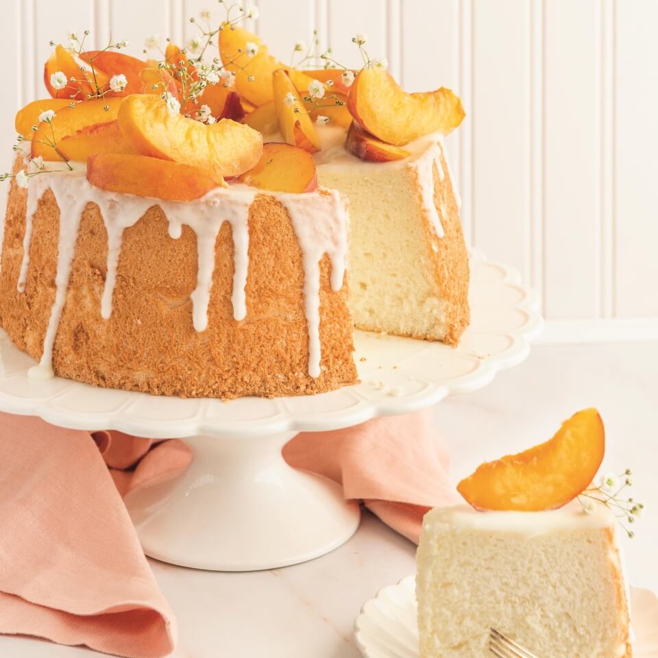 An angel food cake topped with peaches on a white cake stand, with a slice on a plate and light red napkin