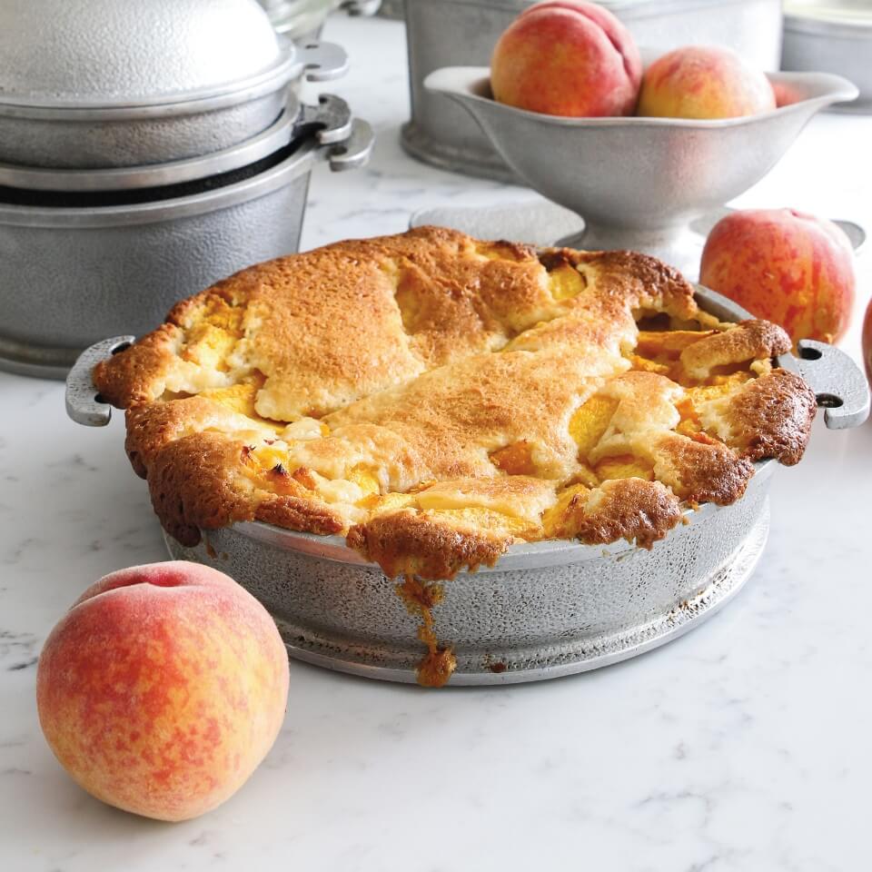 A peach cobbler in a silver dish with peaches around it