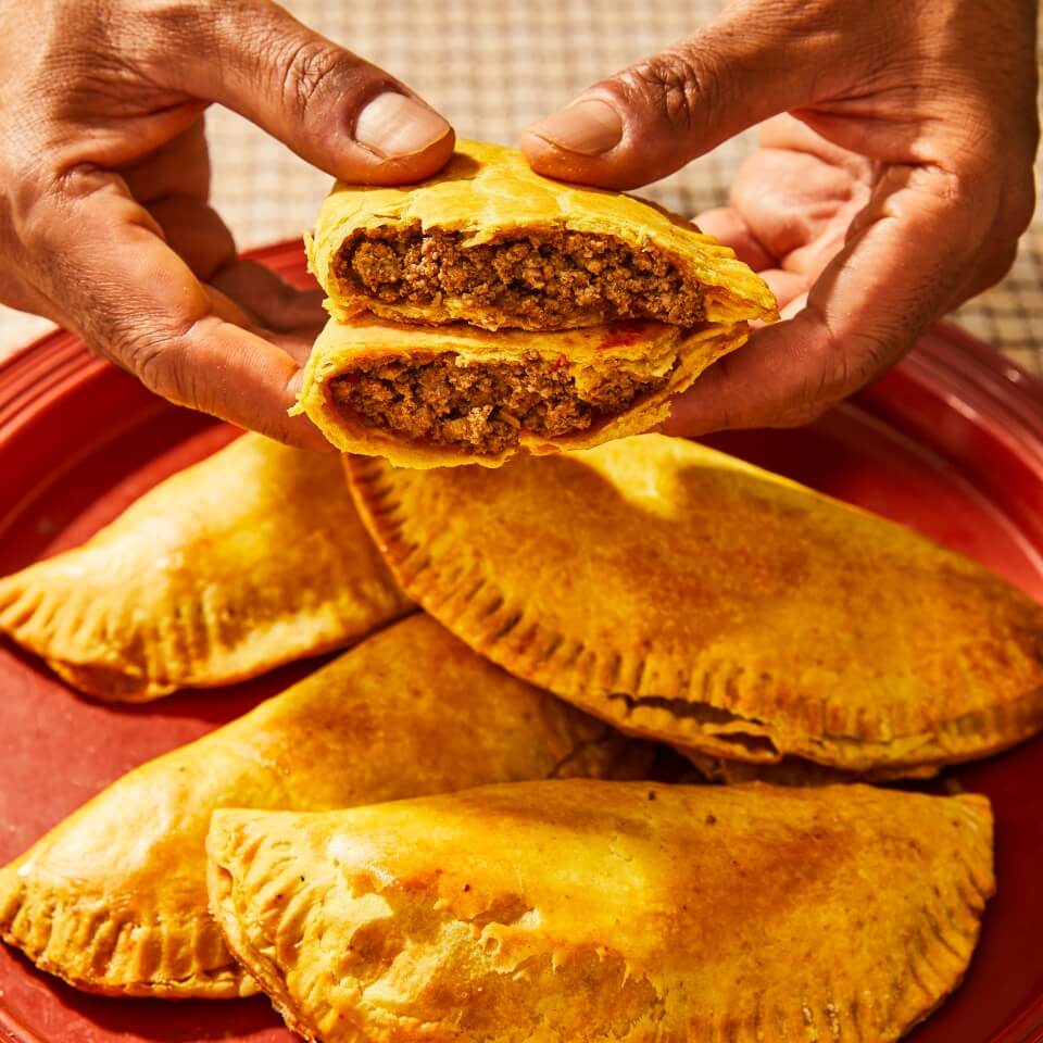 A person's hands holding a Jamaican beef patty broken in half, with more patties visible beneath