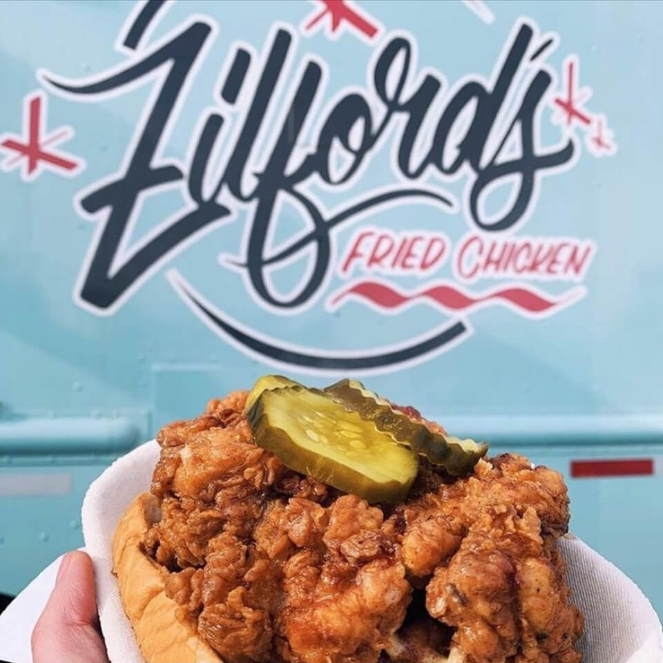 A person holding up fried chicken in front of a blue food truck with a logo reading "Zilford's Fried Chicken"