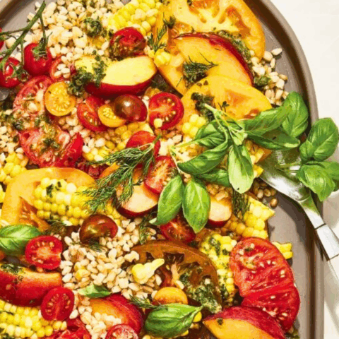 An oval-shaped dish with and heirloom tomato salad with peaches and corn recipe on it and forks next to it