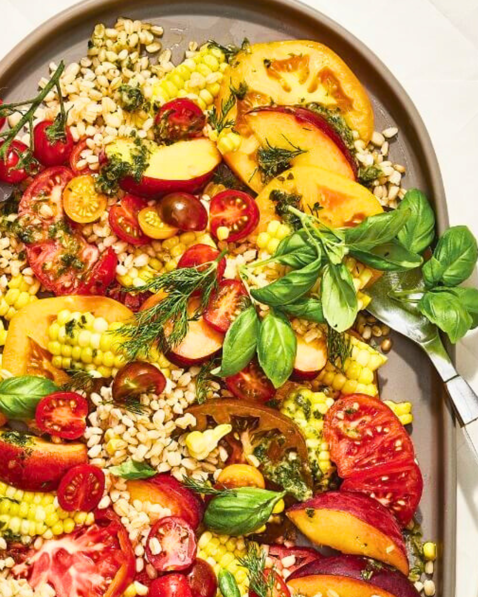 An oval-shaped dish with and heirloom tomato salad with peaches and corn recipe on it and forks next to it