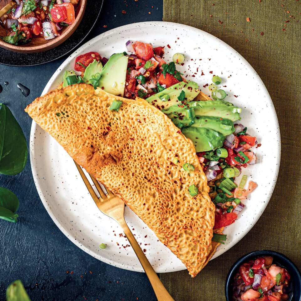 A folded chickpea pancake with a tomato and avocado filling on a white plate with a gold fork. Small bowls of the filling are visible around it