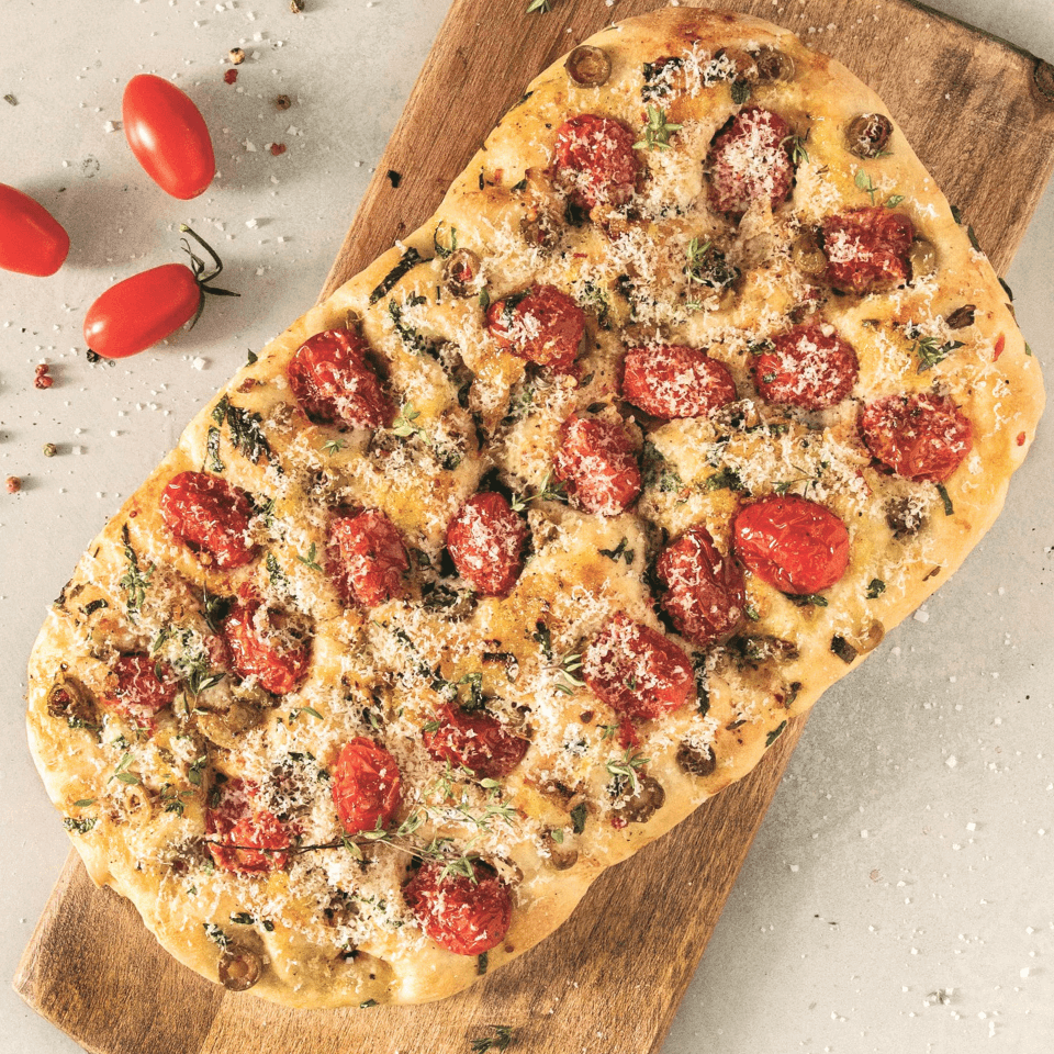 A cherry tomato and green olive focaccia on a wooden serving board, with three whole cherry tomatoes on the counter next to it