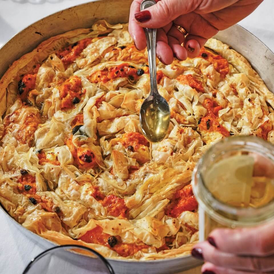 A person drizzling oil with a spoon over a pastry, pumpkin and feta pie