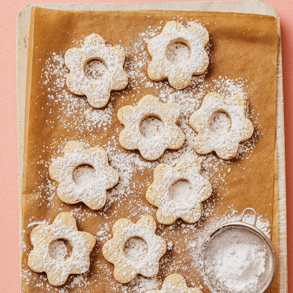 A cookie sheet with flower-shaped cookies and dish of powdered sugar on a pink surface