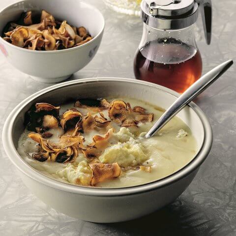 Matty Matheson’s Caramelized Maple Parsnip Soup in a white bowl, topped with golden crispy sunchoke chips, a dollop of ricotta, and a drizzle of maple syrup, garnished with lemon zest and fresh herbs.