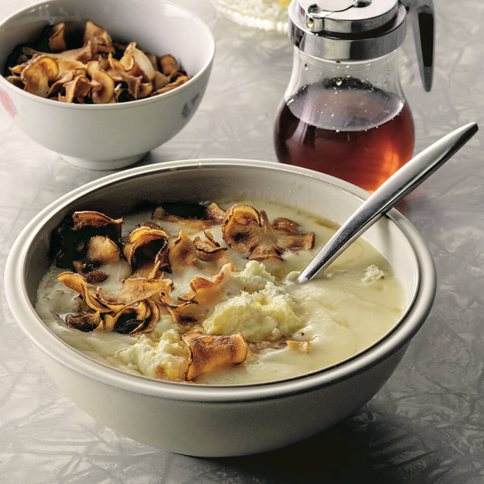 Matty Matheson’s Caramelized Maple Parsnip Soup in a white bowl, topped with golden crispy sunchoke chips, a dollop of ricotta, and a drizzle of maple syrup, garnished with lemon zest and fresh herbs.