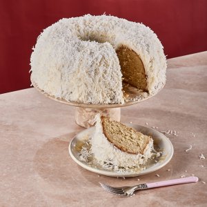 A bundt cake covered with coconut with a slice on a plate next to it on a marble surface with a red background.