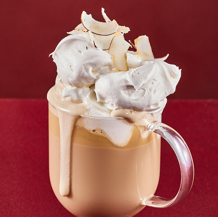 A White chocolate mocha, light brown coffee drink in a clear cup topped with whipped cream and toasted coconut flakes.
