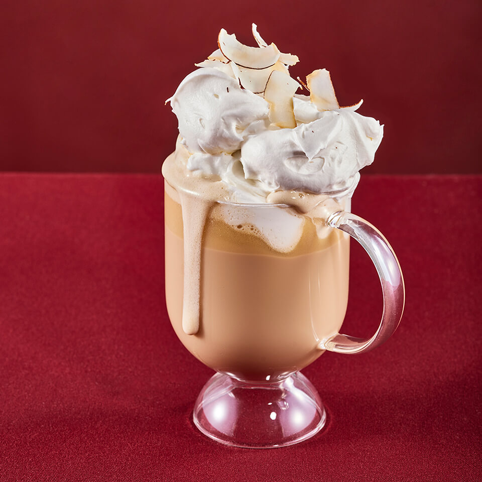 A light brown coffee drink in a clear cup topped with whipped cream and toasted coconut flakes.