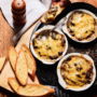 Recipe for Cheesy Mushroom Gratin served in individual white bowls with toasted bread and pepper grinder