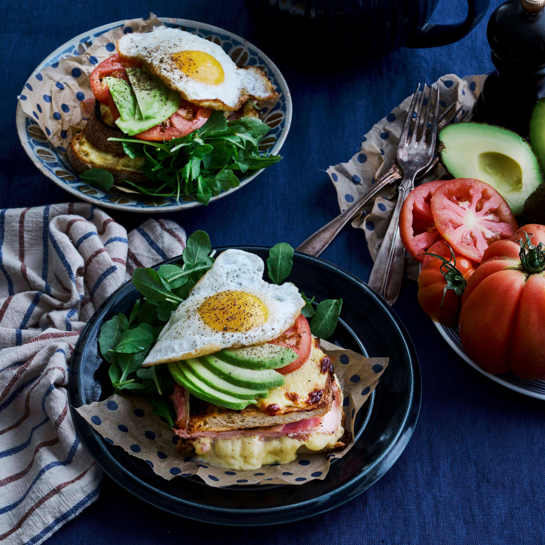 Fried egg layered on top of sliced avocado, tomato and a croque madame sandwich