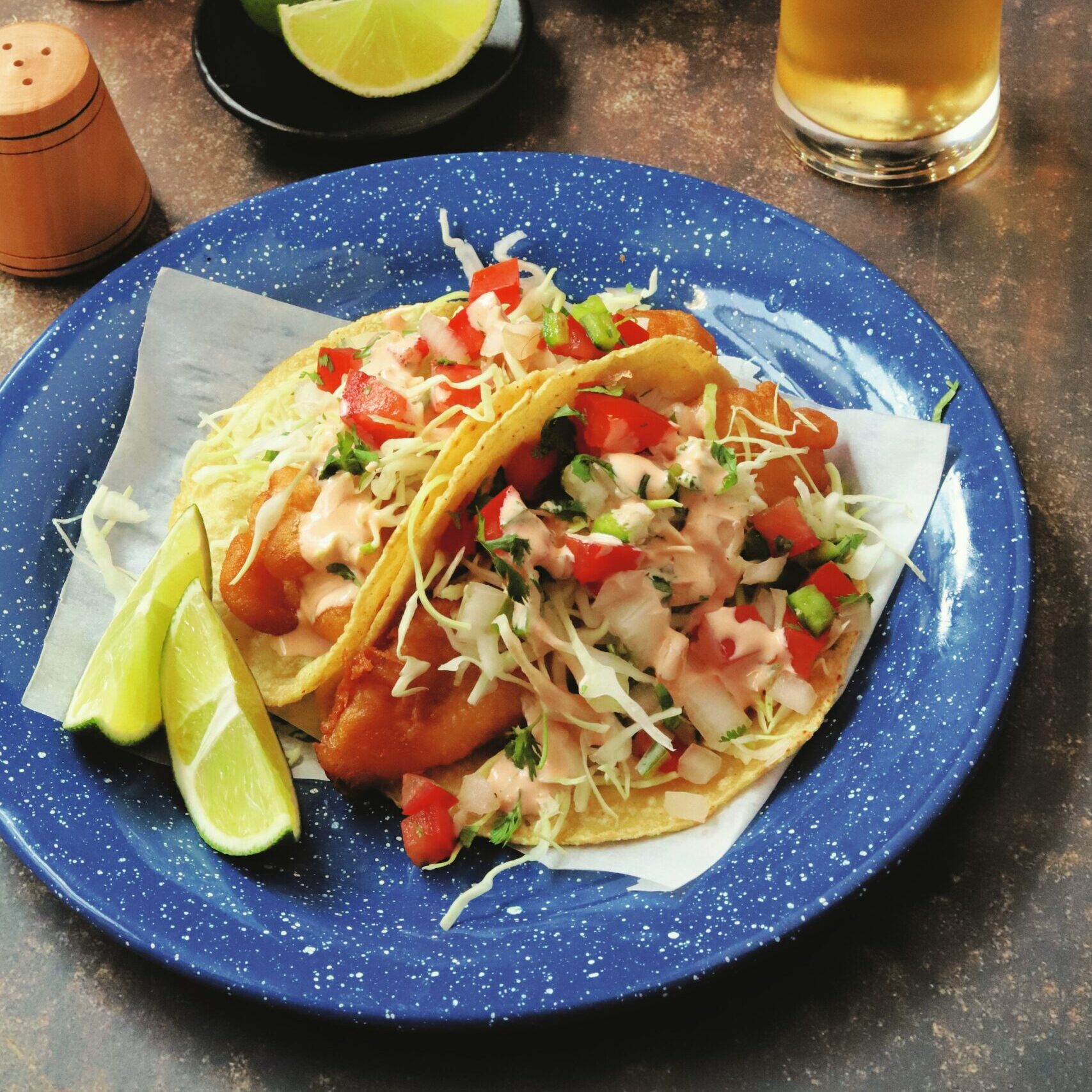 Two baja fish tacos topped with pico de gallo and garnished with two lime wedges.