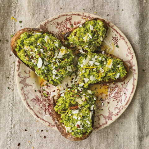 Toast topped with minty peas and shaved pecorino cheese, on a beige decorative plate with burgundy flower design.
