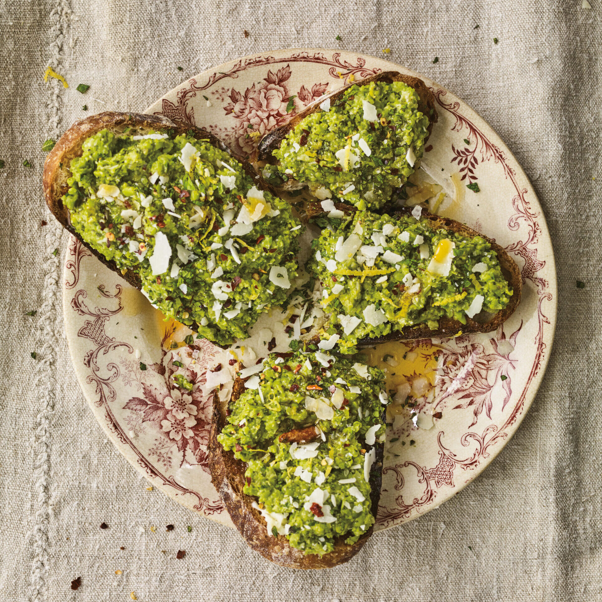 Toast topped with minty peas and shaved pecorino cheese, on a beige decorative plate with burgundy flower design.