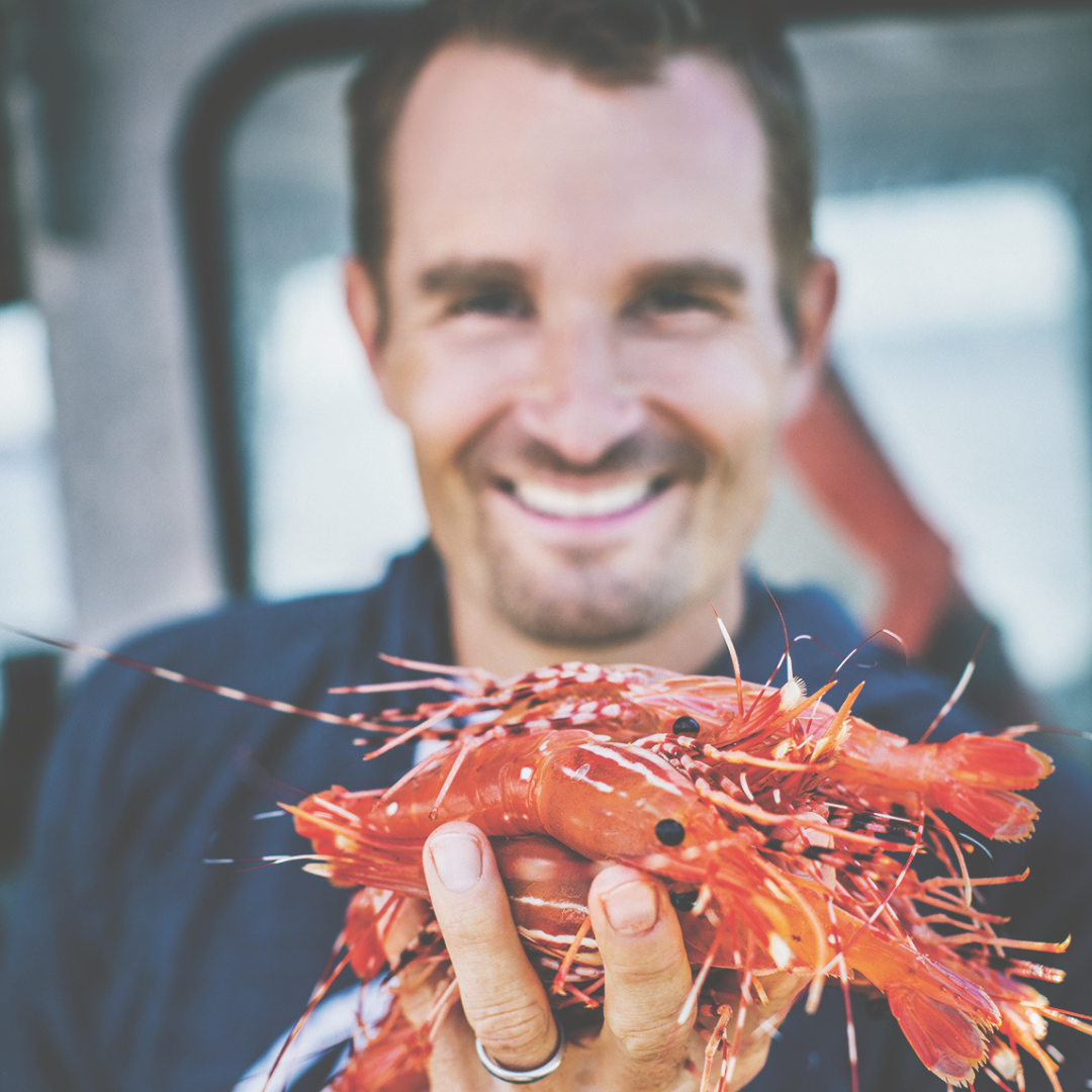 A smiling man holding up a handful of fresh shrimp to the camera.
