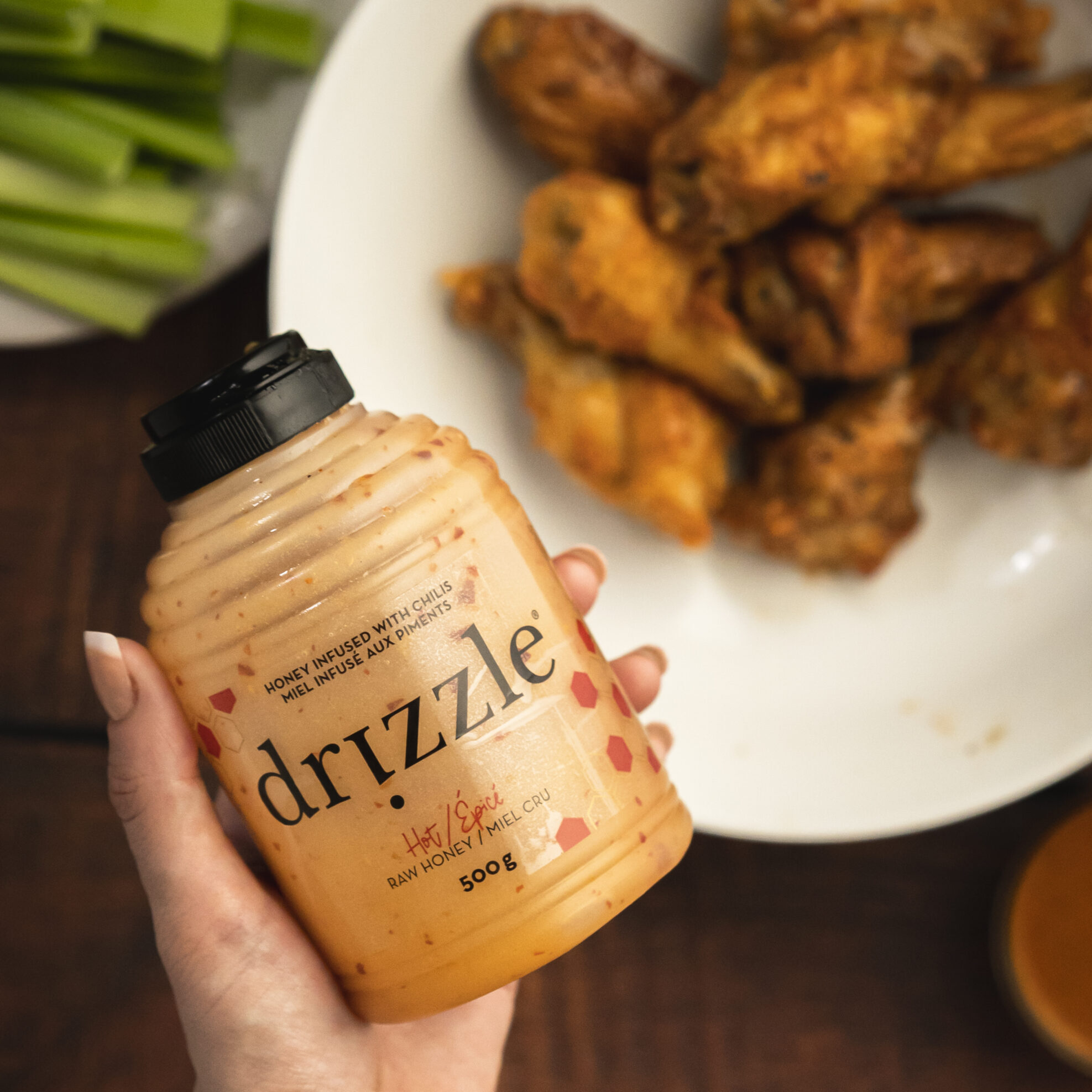 Hand holding a bottle of Drizzle hot honey, with wings in the background.