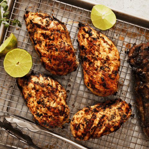 Four grilled chicken breasts on a wire rack with lime halves next to it.