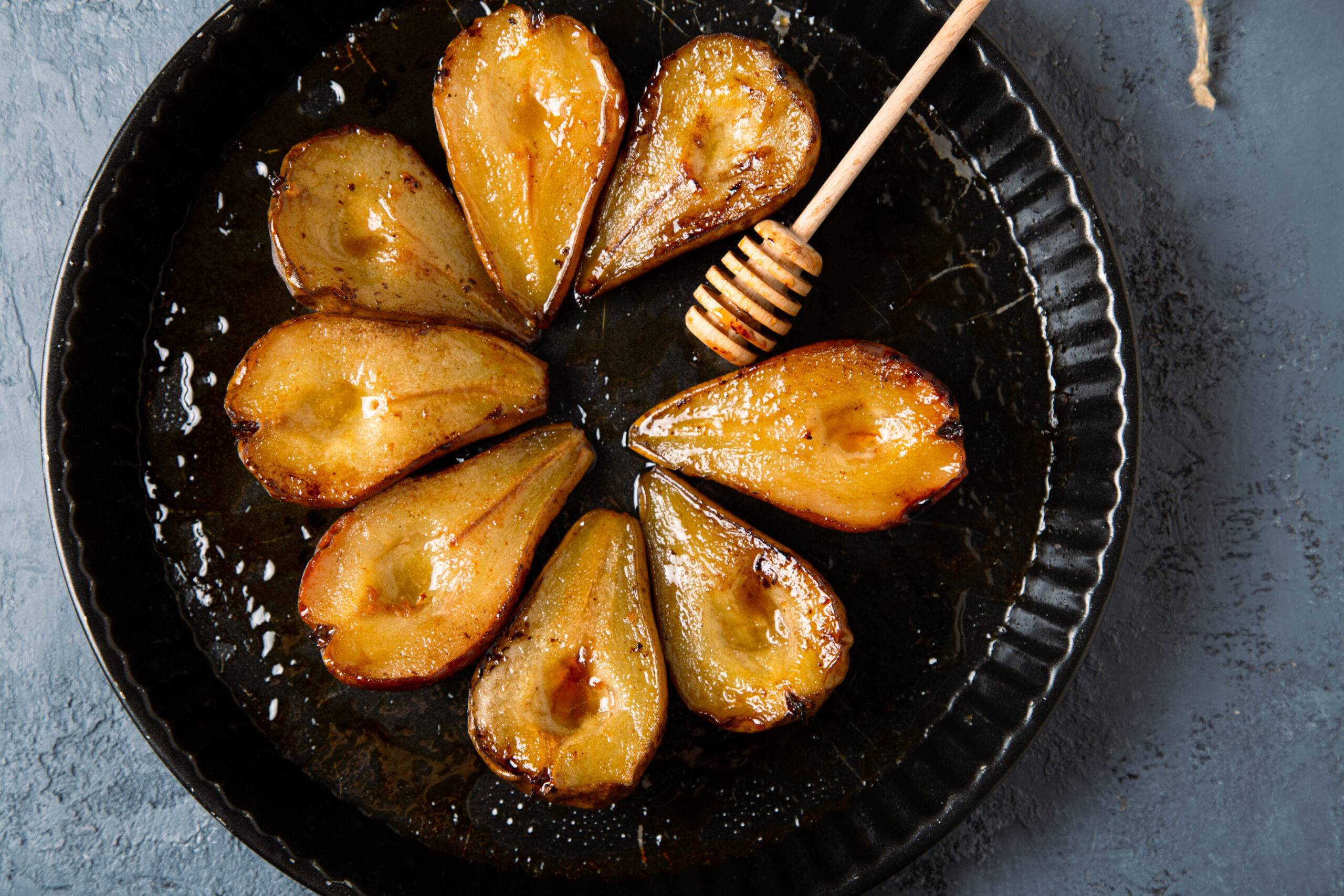 Baked pears with honey, Modena vinegard and cinnamon, and honey dipper in a round baking tray on a grayish blue cocrete background