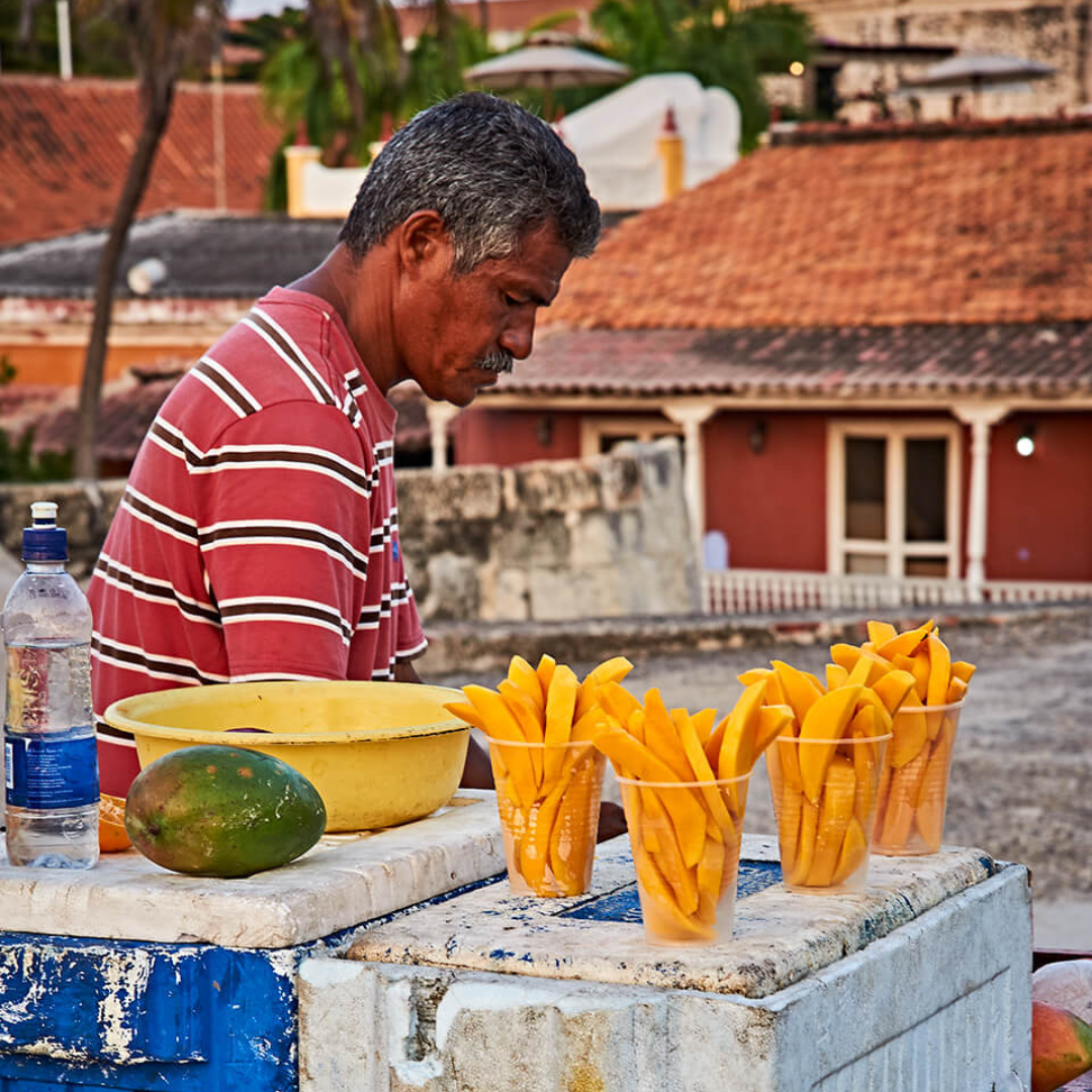 An old man in a red striped shirt with slices of mango in plastic cups on top of the vendor stand in front of him.