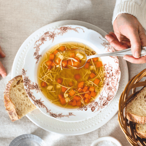 One hand holding a spoon in a bowl of Anna Olsen's chicken noodle soup, with a piece of bread next to it.