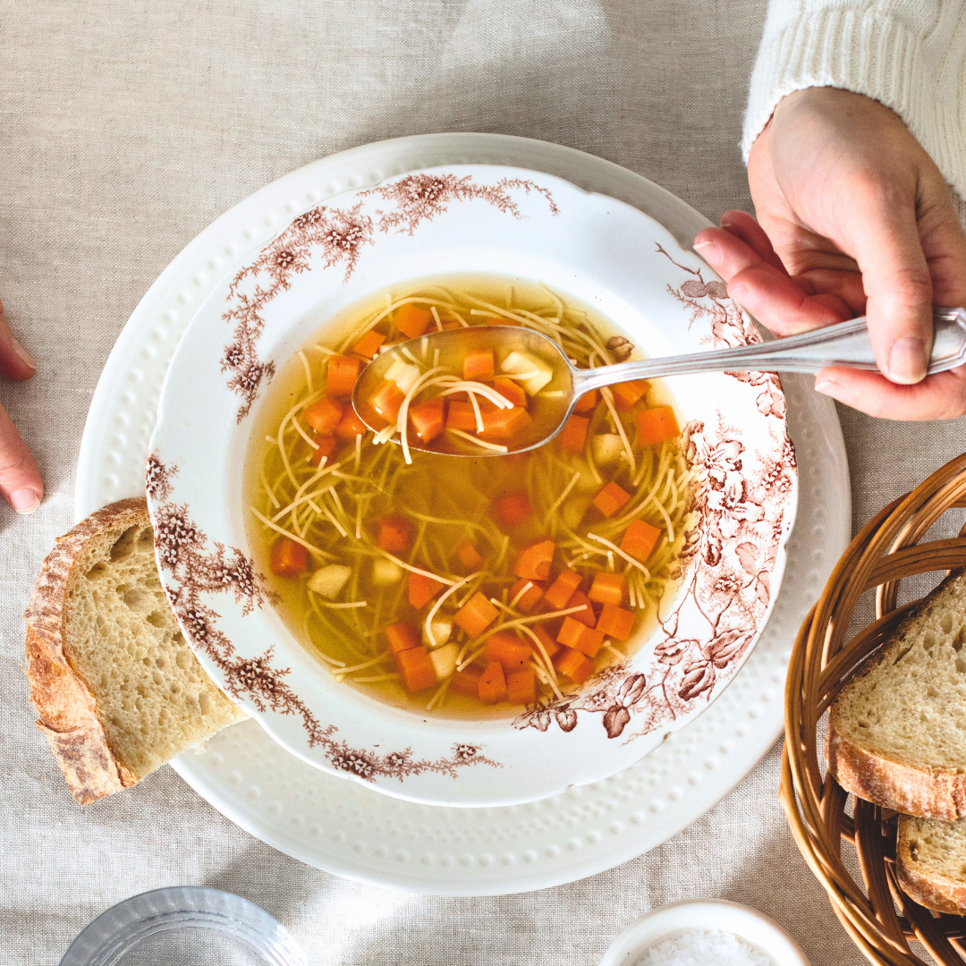 One hand holding a spoon in a bowl of Anna Olsen's chicken noodle soup, with a piece of bread next to it.